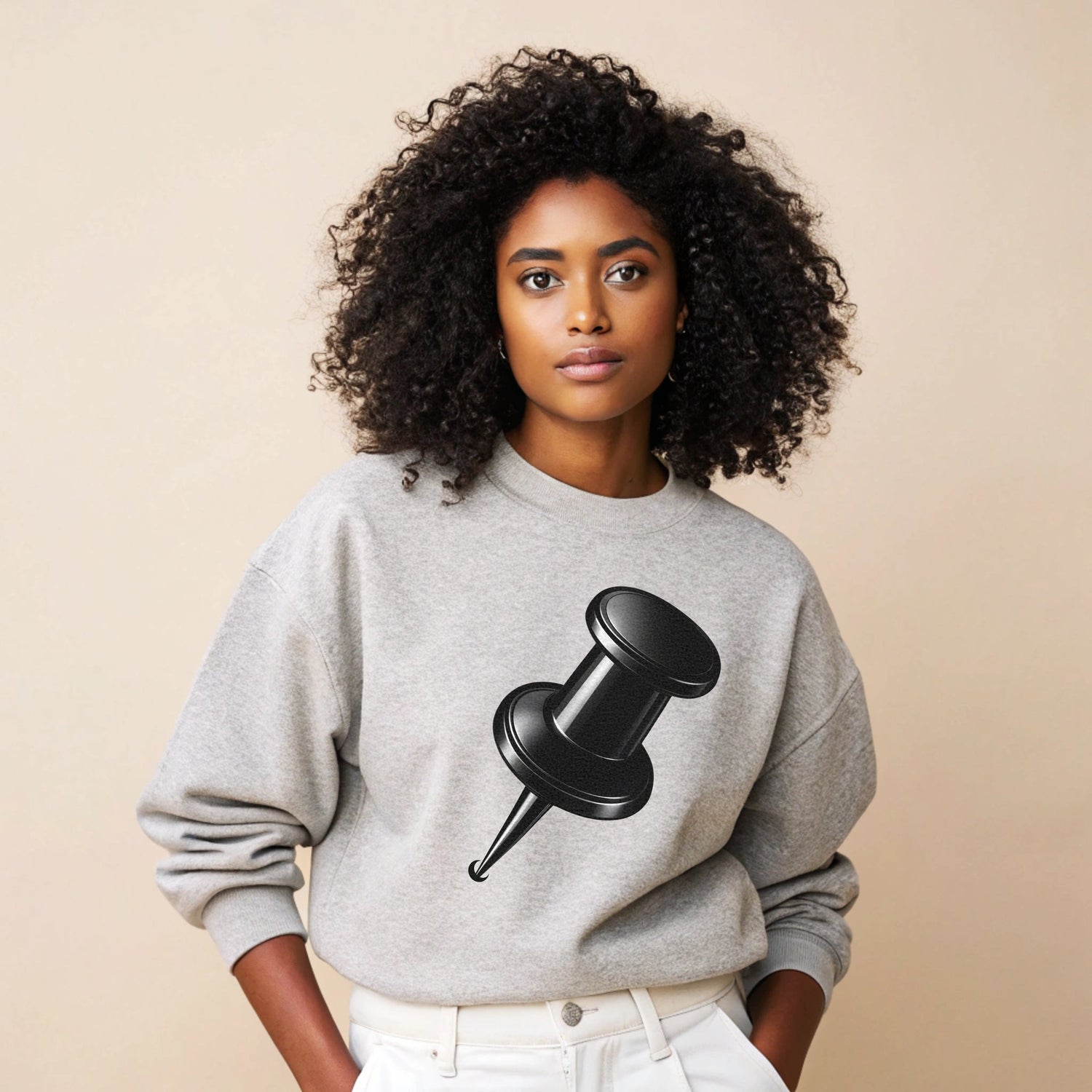 Black woman with natural curly hair wearing a grey oversized sweatshirt with a black pin graphic, paired with white trousers, standing against a soft beige background.
