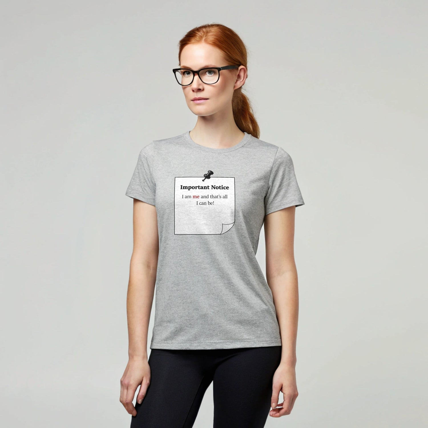 A woman in black leggings wearing a grey classic cotton t-shirt featuring a black-outlined square note with a black push pin. The note displays the slogan: 'Important Notice: I am me and that’s all I can be,' with 'me' in bold red for emphasis.